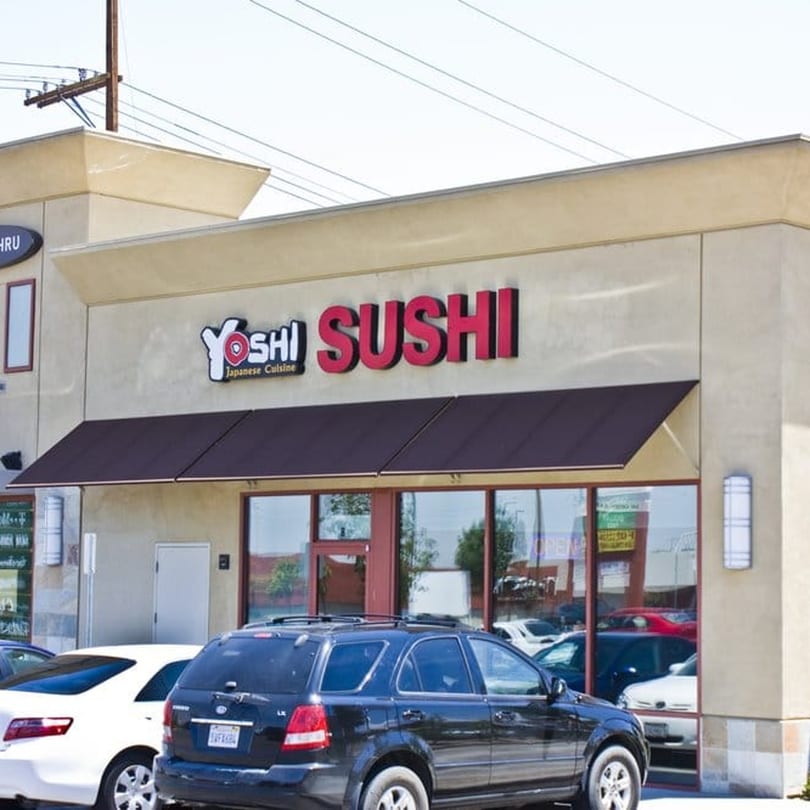 Exterior of Yoshi Sushi restaurant with a red sign and dark awnings, cars parked in front.